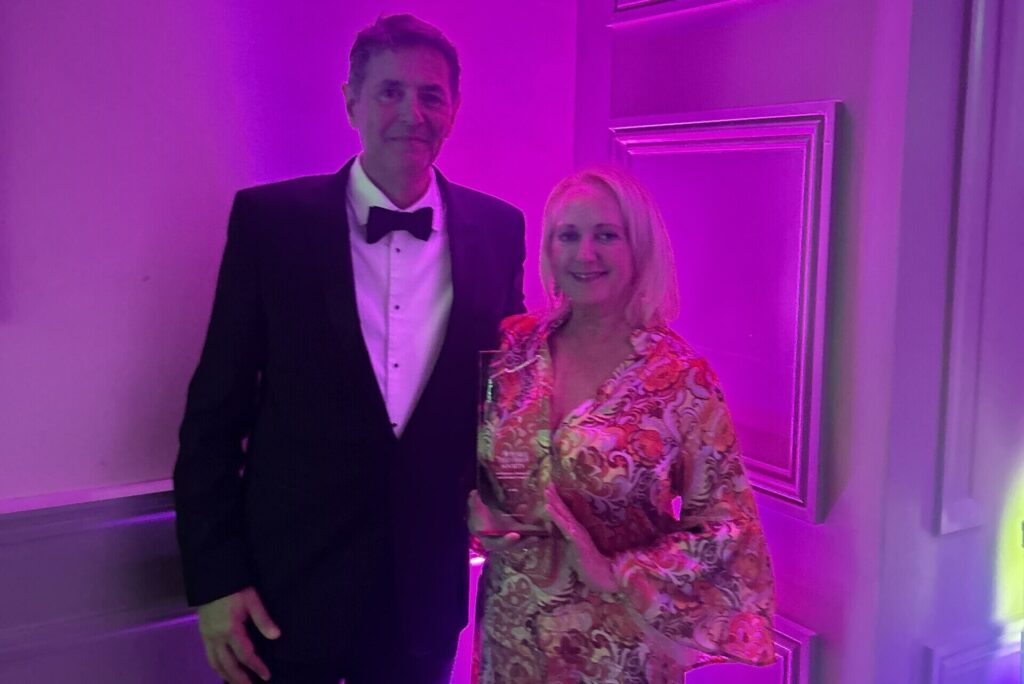 Man in tuxedo and woman in patterned dress holding an award under purple lighting